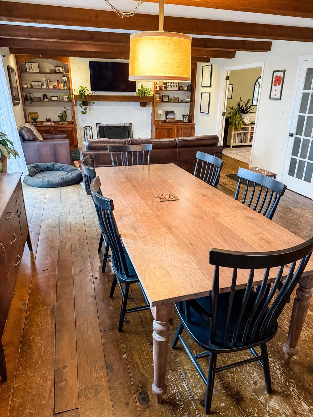 May include: A wooden dining table with six black chairs in a well-lit room. The table has a light wood surface and is surrounded by chairs with curved backs. The room features a fireplace, a leather sofa, and a large light fixture.